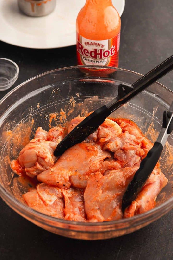 Photo of buffalo chicken thighs marinating in a large glass bowl. We a pair of tongs resting on the side of the bowl.